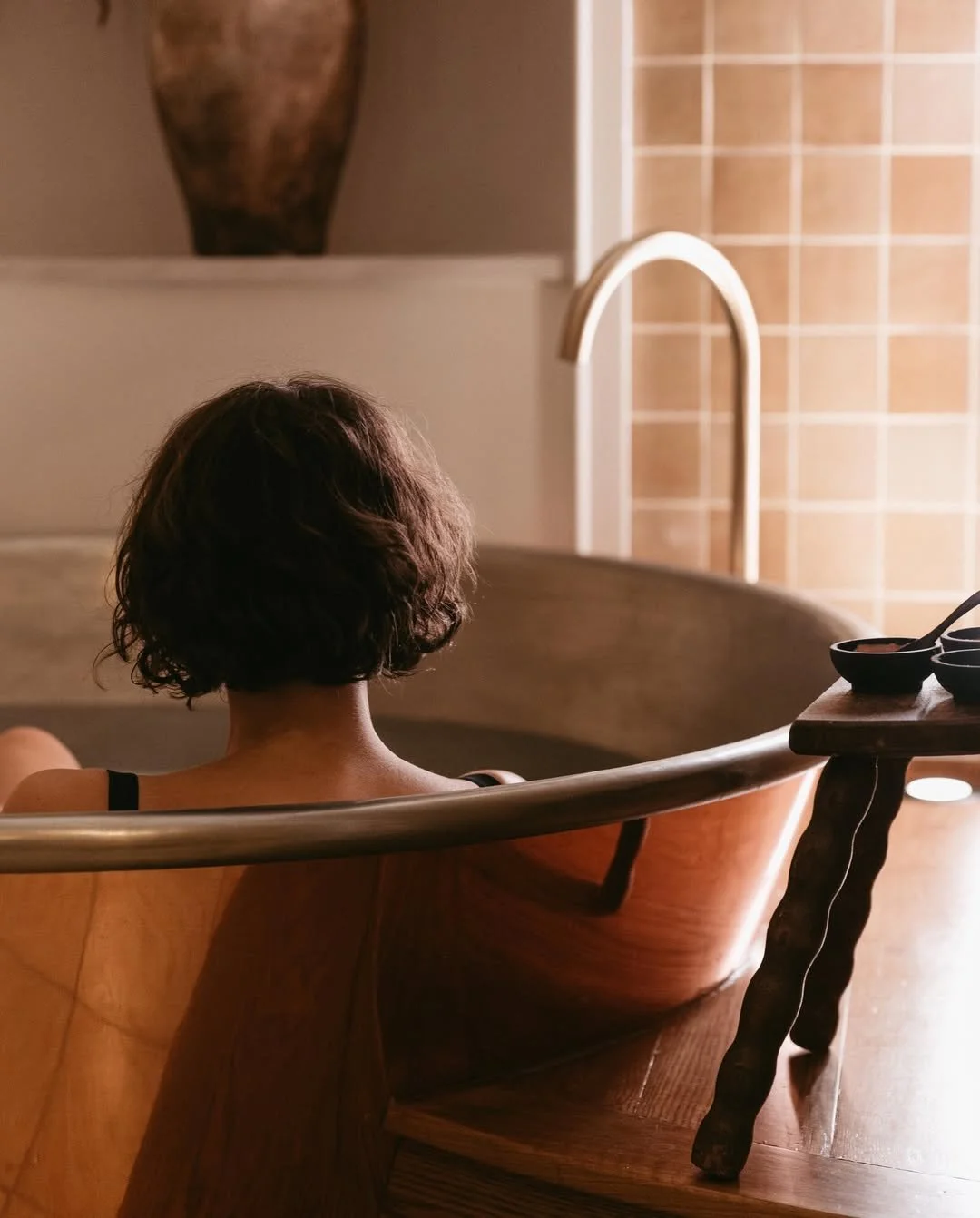 A person with short, curly dark hair sitting in a wooden bathtub, viewed from behind, with a towel on their shoulder and a black strap on their arm.