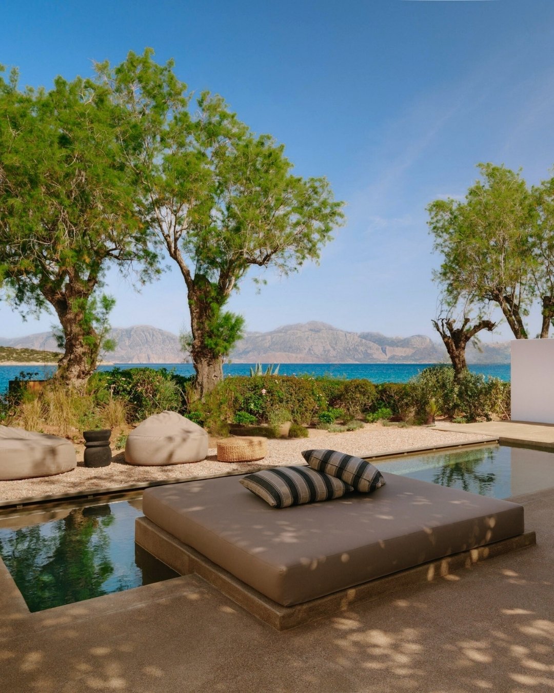 Outdoor seating area with cushioned lounge bed and striped pillows, overlooking trees, shrubs, and mountains near the water on a sunny day.