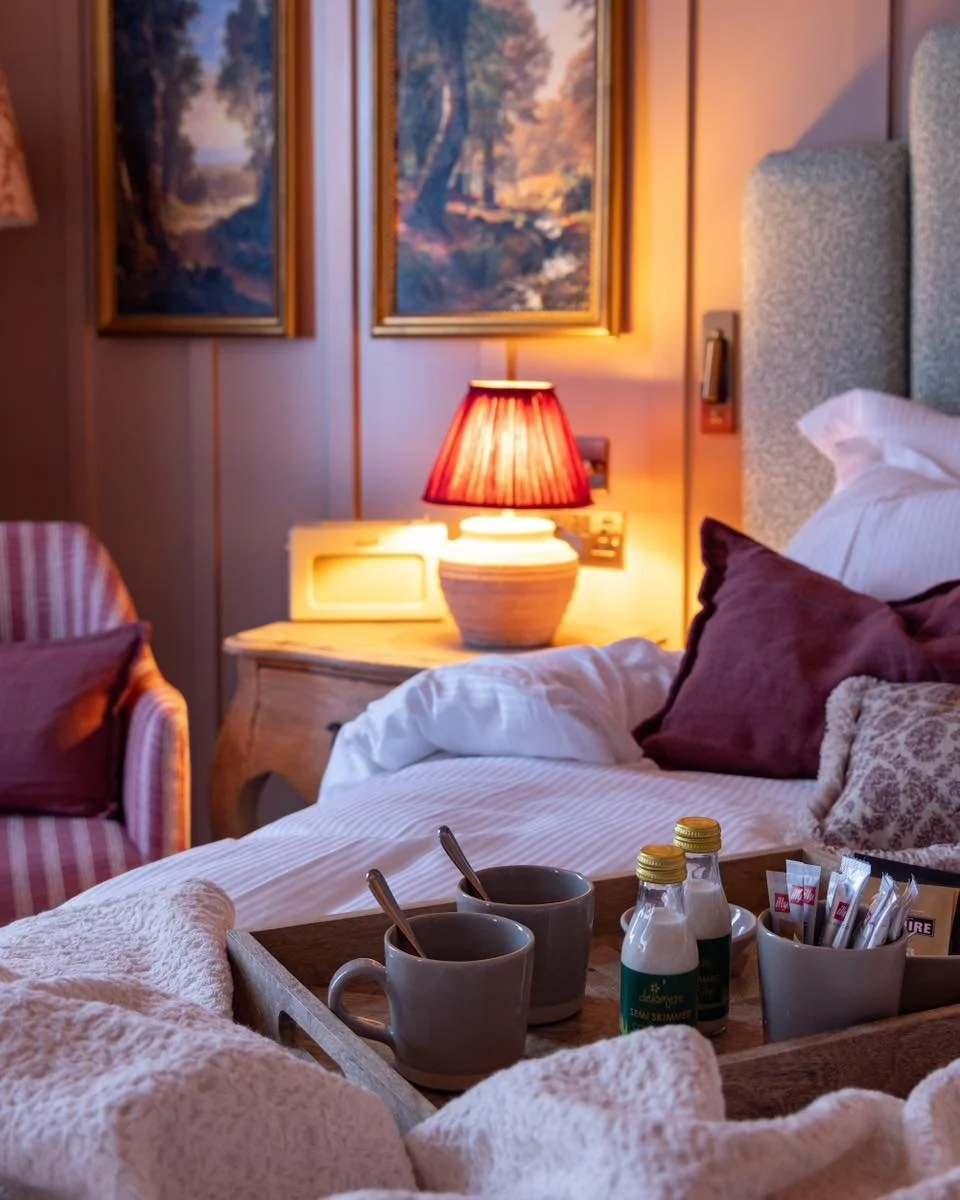 Cozy hotel room with a bed, pillows, bedside table with a red lamp, artwork on the wall, and a tray holding coffee mugs, bottles, and condiments.