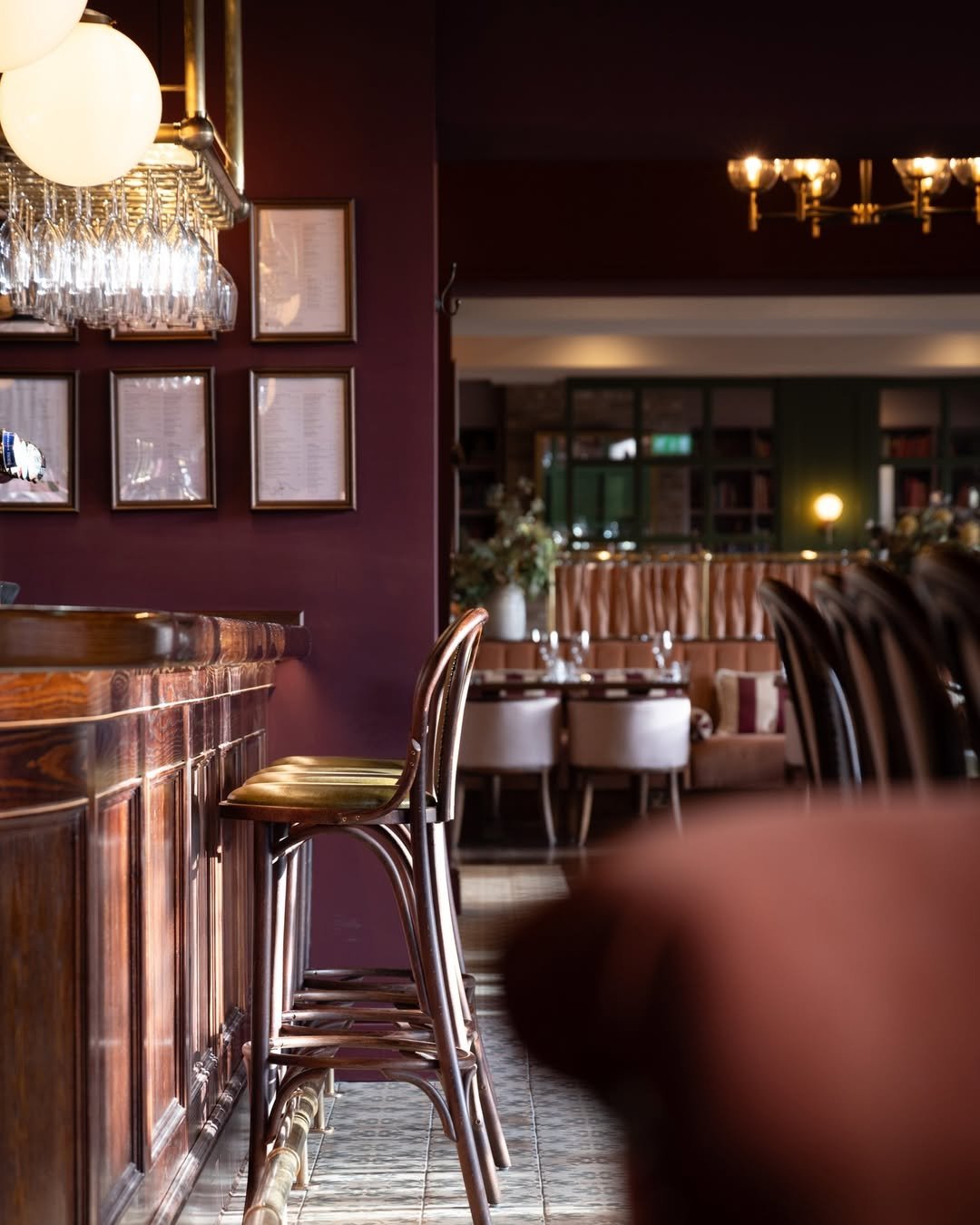 An interior of a restaurant or bar with a wooden counter and high chairs, maroon walls, framed pictures, and a cozy dining area with tables, chairs, and floral decorations.
