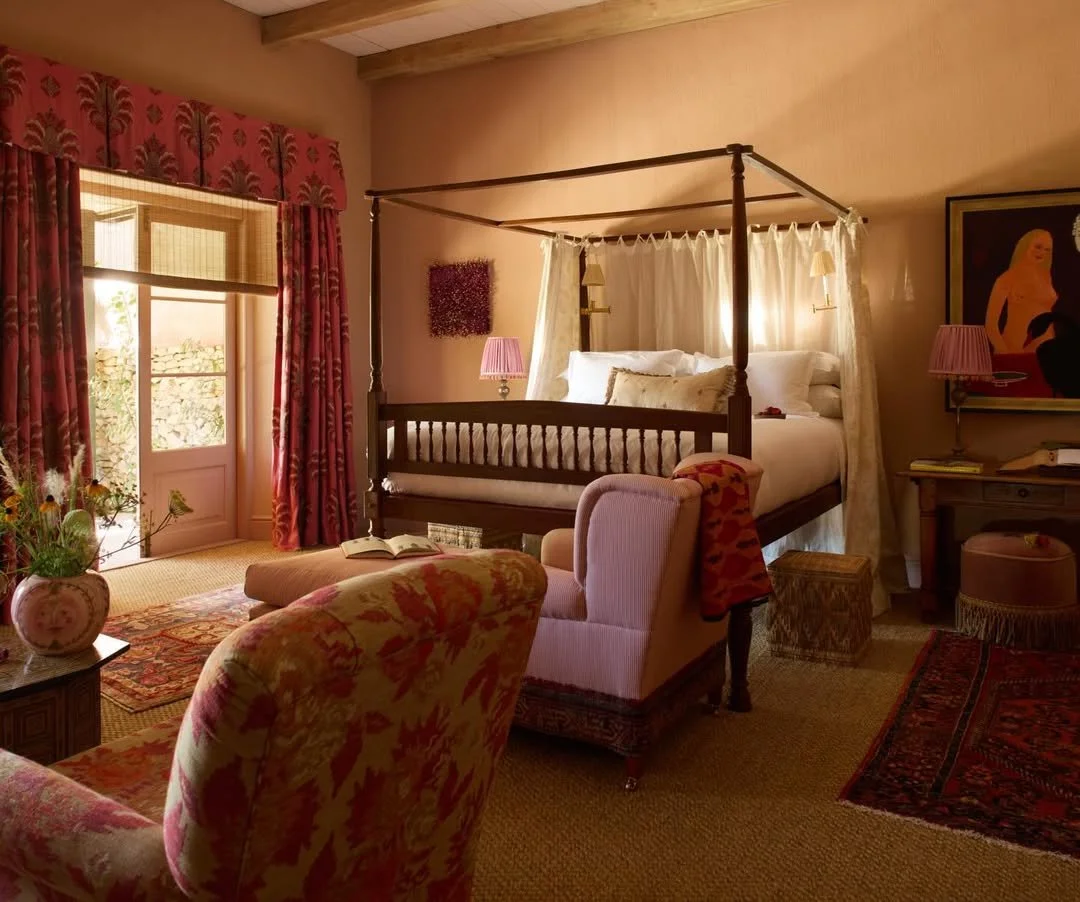 A cozy bedroom with a four-poster bed draped in white curtains, pink walls, floral curtains, and vintage furniture, including armchairs, a side table, and a desk with lamps.