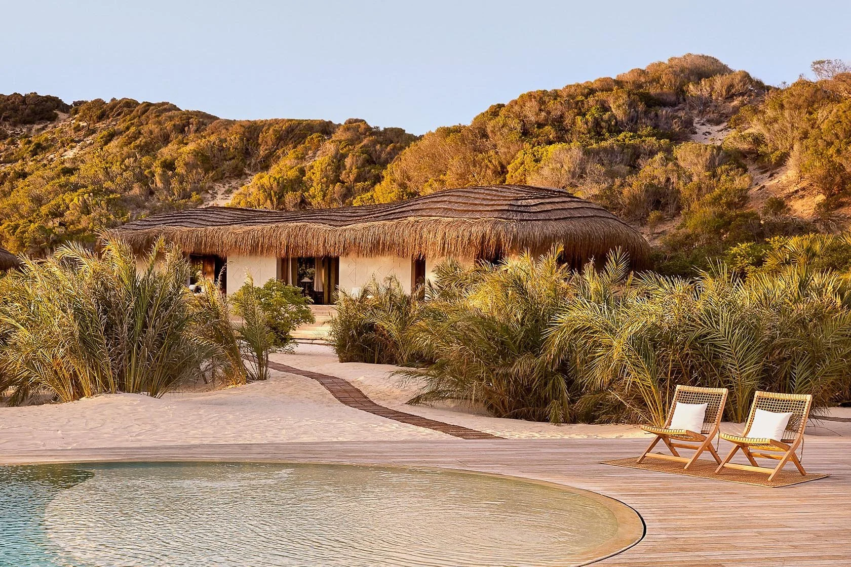 Beachside bungalow with thatched roof, surrounded by palm bushes, in a sandy area with a swimming pool, wooden deck, and two lounge chairs, with hillside in the background.