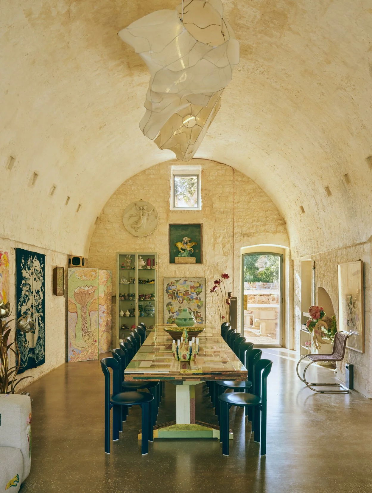 A long wooden dining table with black chairs in a rustic, stone-walled room with artwork, a glass display cabinet, and large windows letting in natural light.