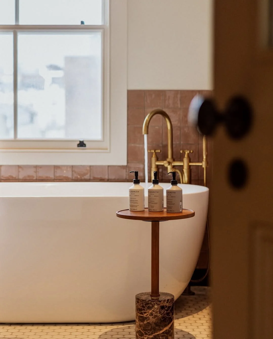A white bathtub with gold faucet fixtures next to a window with frosted glass, a small wooden stand with three bottles of skincare products, and brown tiles in a bathroom.
