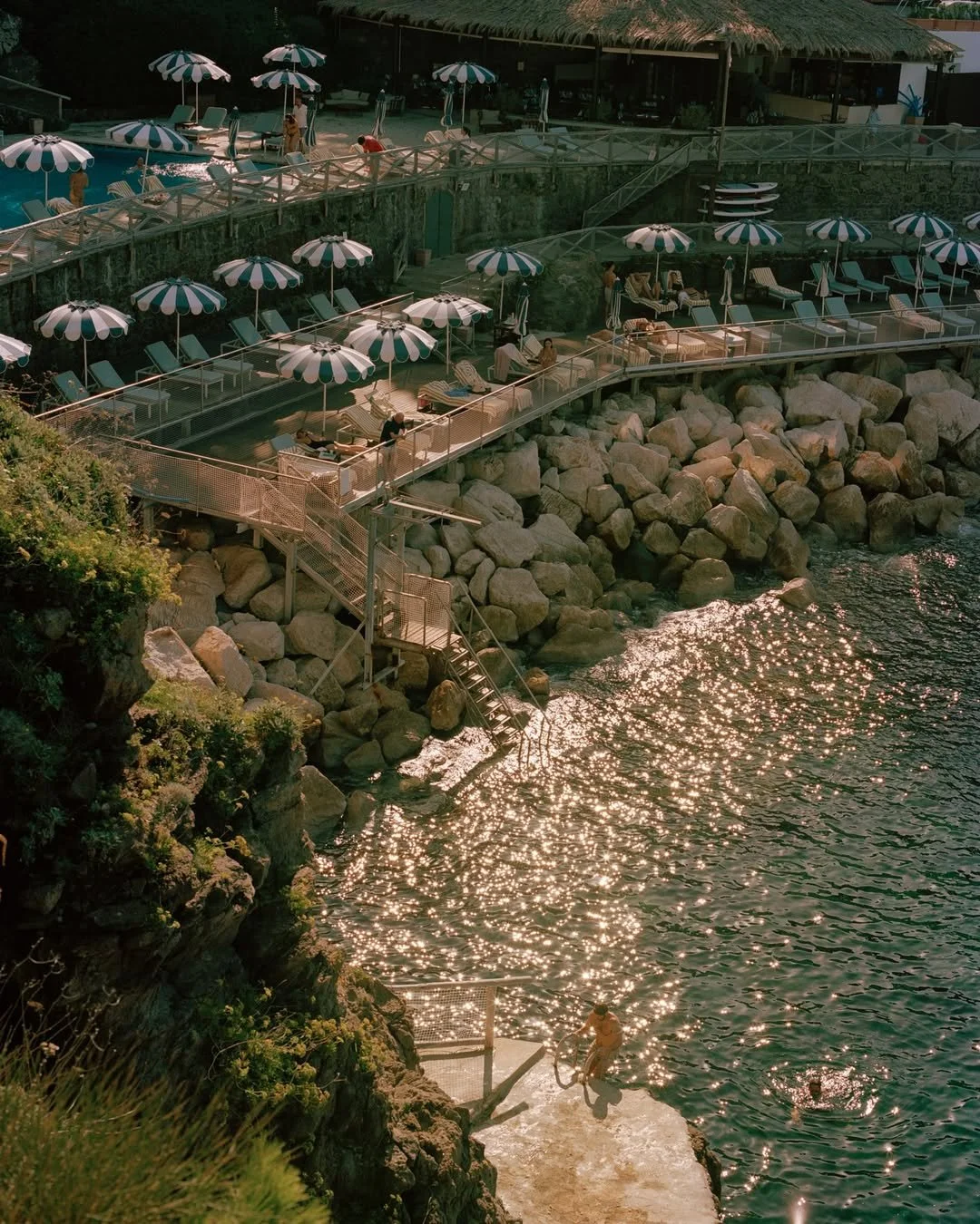 A multi-level seaside lounge area with striped umbrellas, lounge chairs, and people relaxing by the water and swimming. Rocky shoreline and staircases leading down to the water.