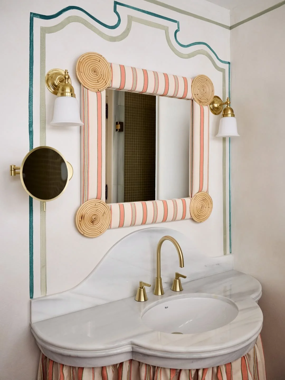 Bathroom vanity with marble countertop, gold fixtures, and a mirror framed with striped fabric and decorative rope details. Wall-mounted lights and a small magnifying mirror are also present.