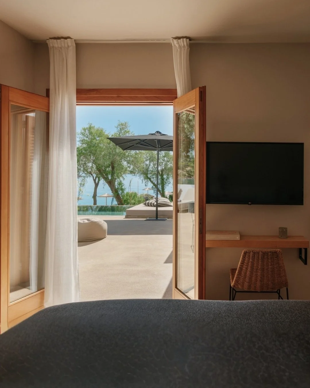 A view from inside a room looking out through a glass door to an outdoor patio with a pool, umbrella, and trees overlooking a body of water.