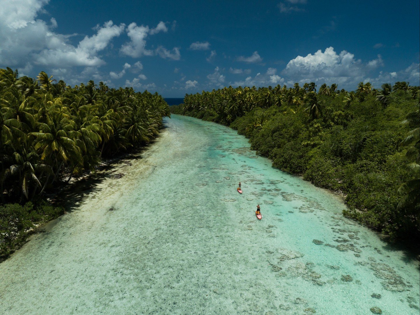 Two people paddleboarding on a clear, shallow waterway surrounded by dense green tropical vegetation under a partly cloudy sky.