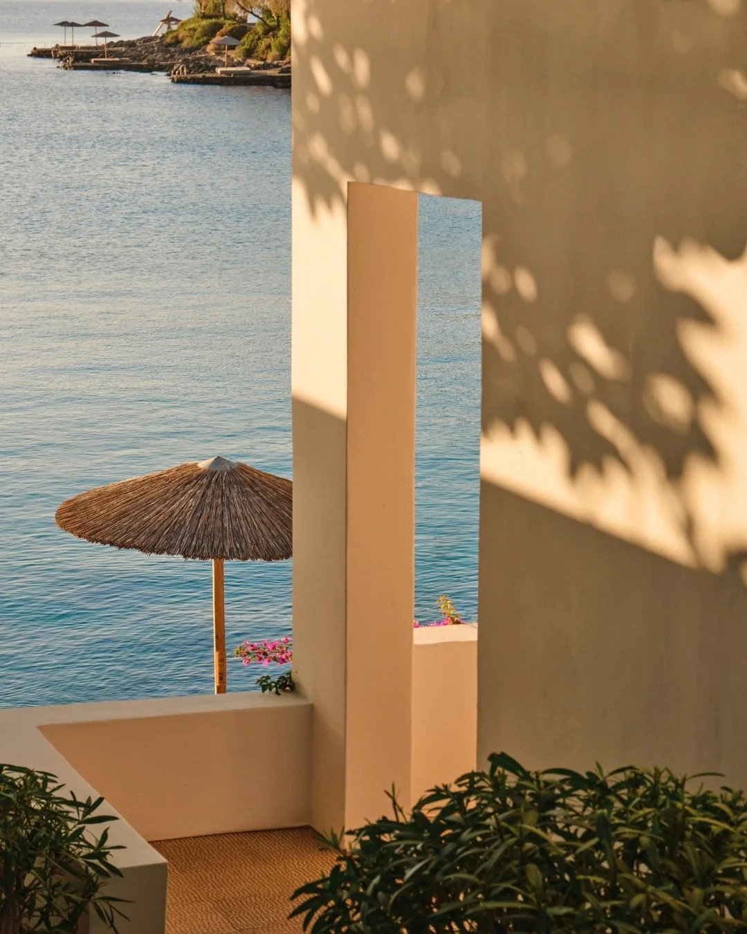 View of a beach scene through a window with a white frame, featuring a thatched umbrella, water, and distant shoreline with umbrellas and greenery.