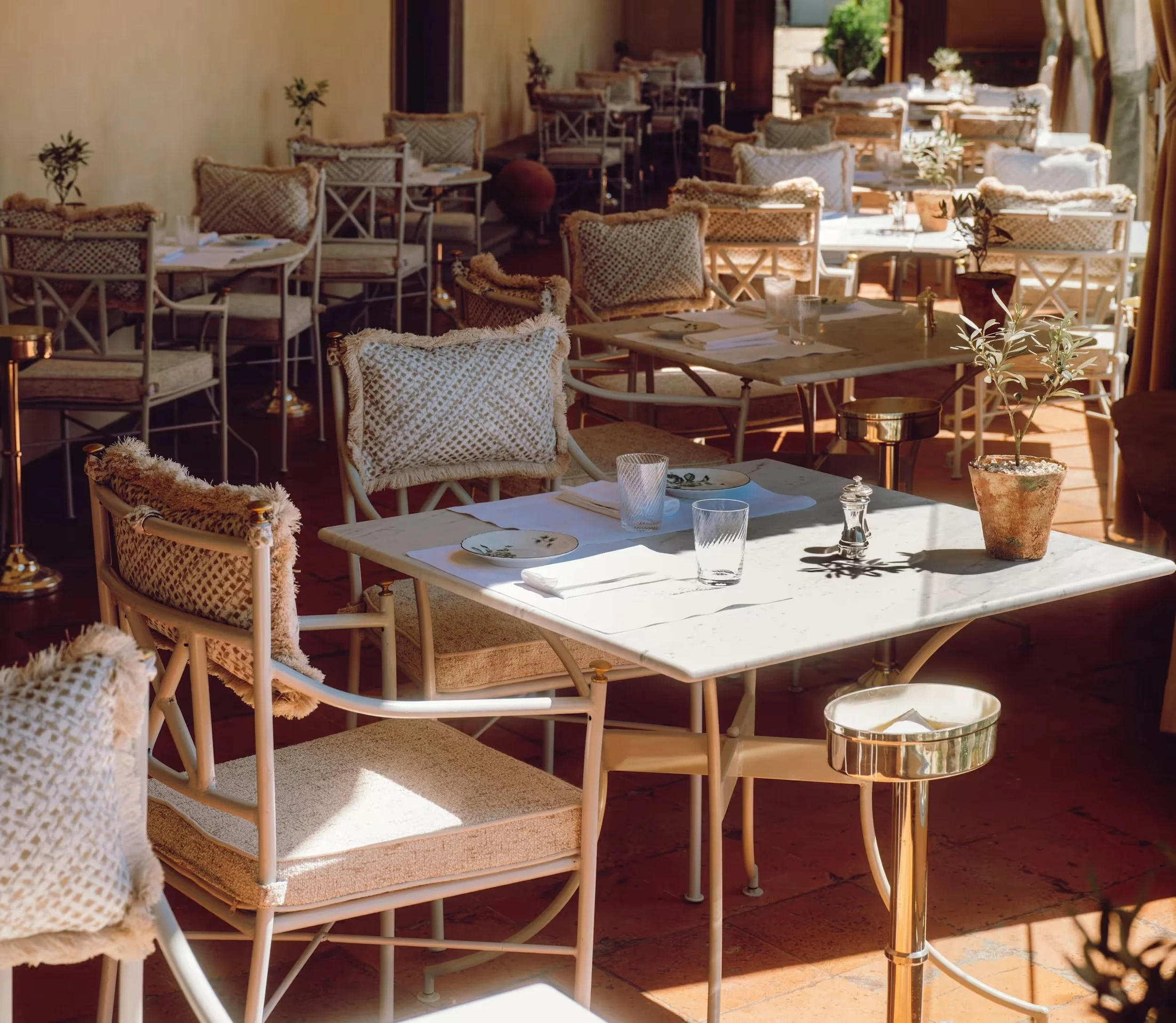 There are white tables set up for a meal, with plates, glasses, and napkins, and wooden chairs with woven cushions arranged around the tables. Sunlight is streaming in, creating a warm and inviting atmosphere, with some plants placed on the tables and a golden side table nearby.