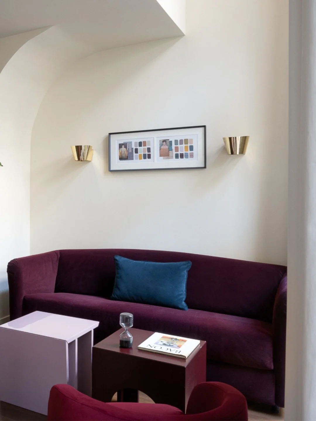 Living room with a burgundy velvet sofa, a blue pillow, a side table with a magazine, a small hourglass, and a framed artwork on the wall with two photographs and color swatches, flanked by gold wall sconces.