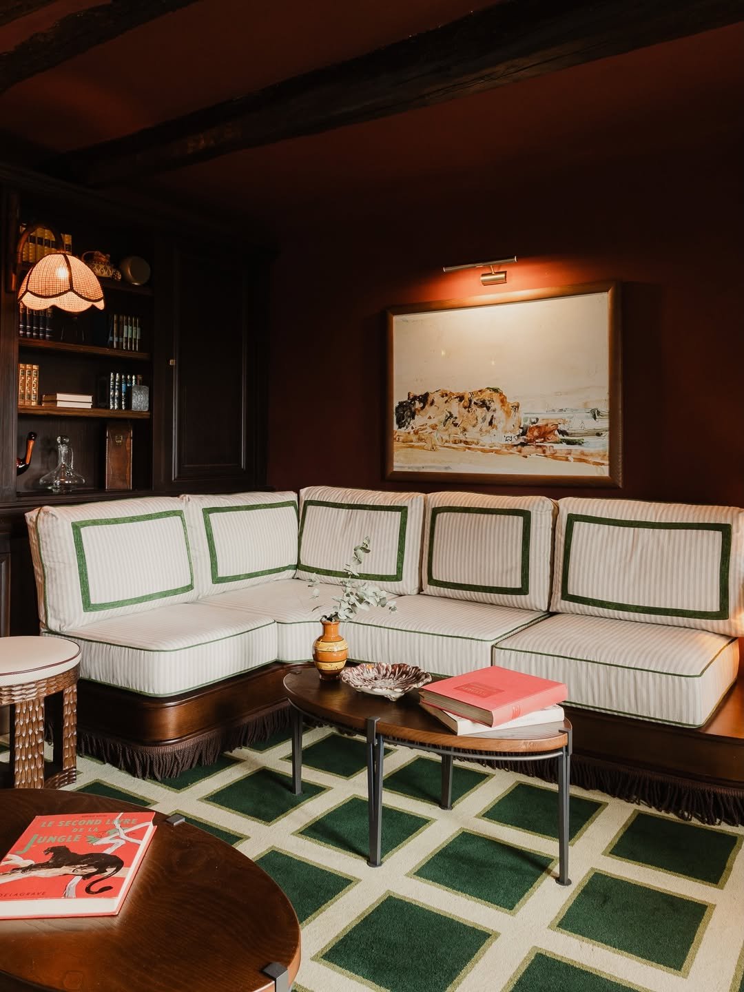 Living room with white sofa with green trim, dark wood bookshelves, a framed painting of a coastal landscape, a small wooden coffee table with a vase and books, a patterned green and white rug, and a side table with a book and a magazine.