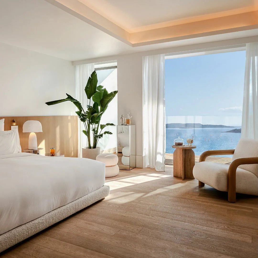 A bright, modern bedroom with a large bed, a wooden headboard, a bedside table with a lamp, a large green plant, a white armchair, a small side table, and a view of the ocean through large windows with white curtains.
