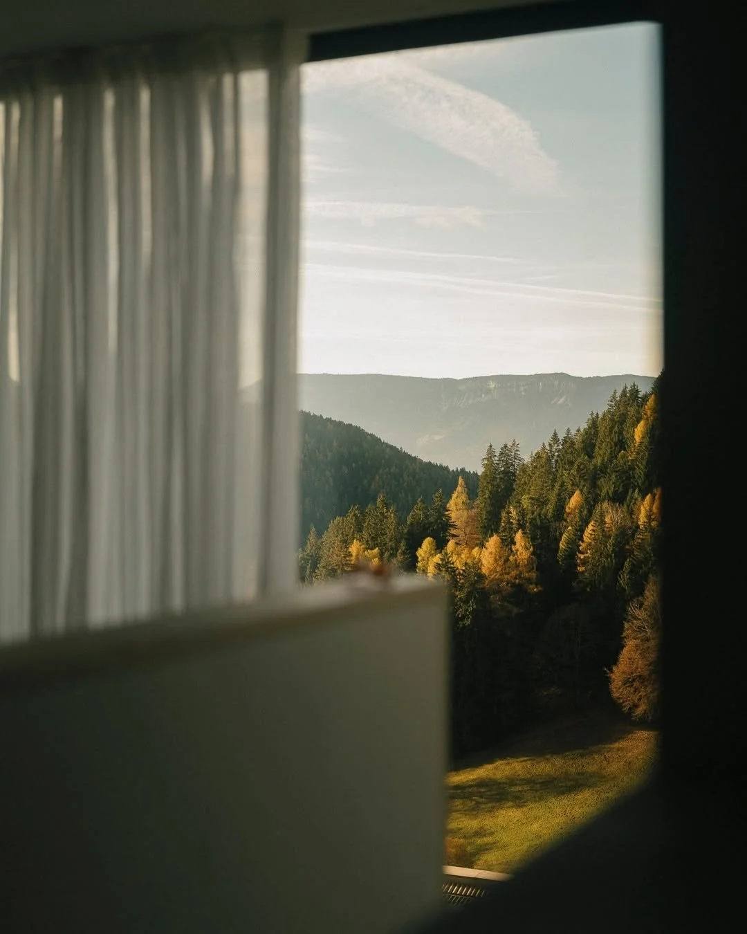 View of a mountain landscape with green trees, seen through a partially open window with white curtains.