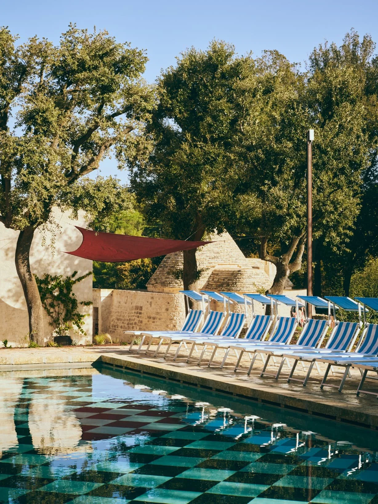 Swimming pool area with striped lounge chairs, blue umbrellas, trees, and a shade sail, near a stone building under a clear blue sky.