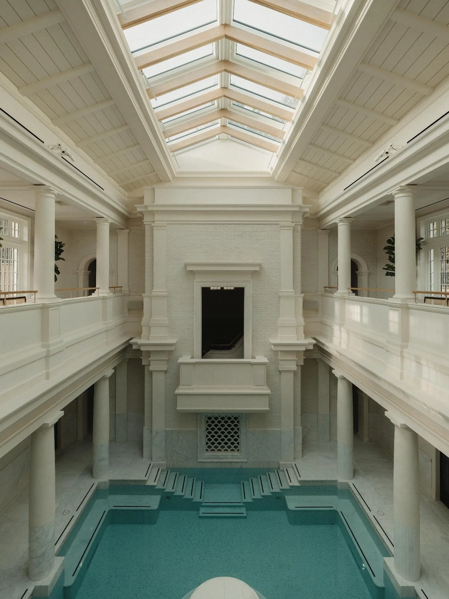 Interior of a luxurious building with a large skylight ceiling, white walls, columns, and a swimming pool below.