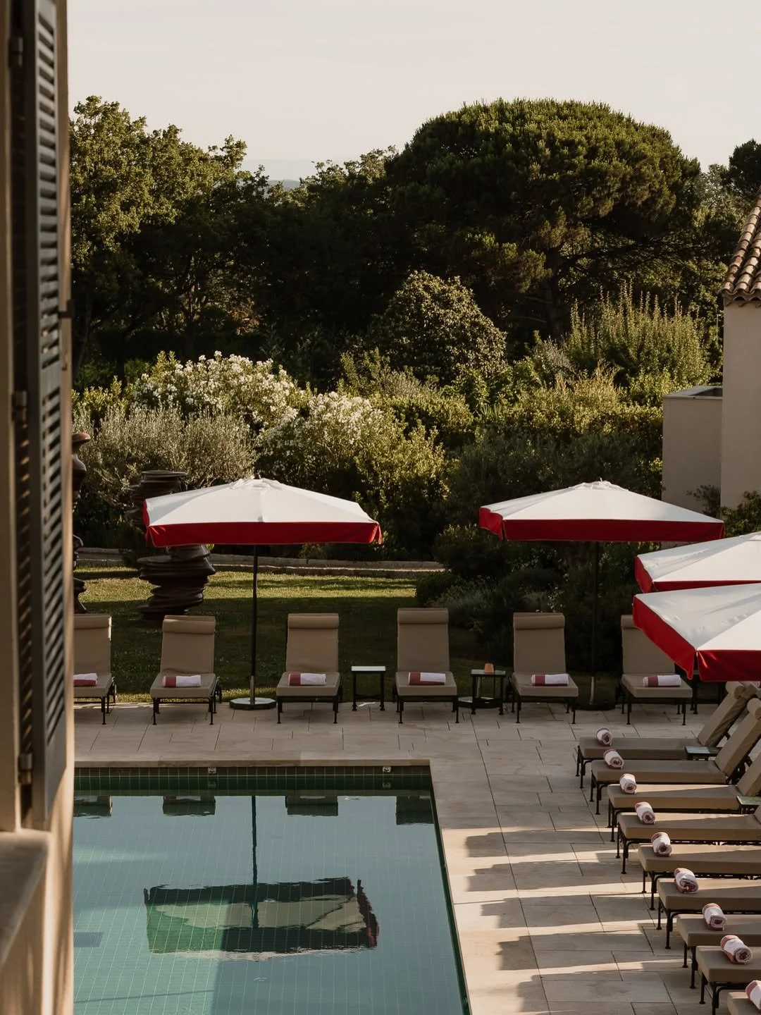 Poolside area with lounge chairs, rolled towels, and red and white umbrellas, surrounded by greenery
