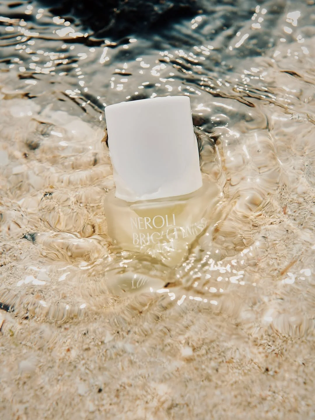A bottle of Neroli Brightening Milk is floating in shallow water with ripples and sunlight reflections.
