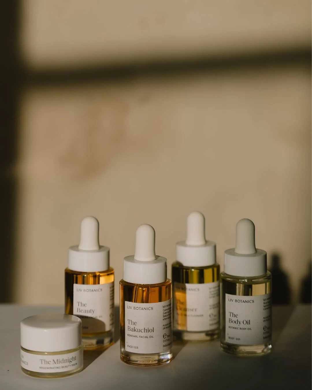 A collection of skincare products including bottles of facial and body oils, with labels such as The Midnight, The Beauty, The Bakuchiol, and The Body Oil, arranged on a surface with a neutral background.