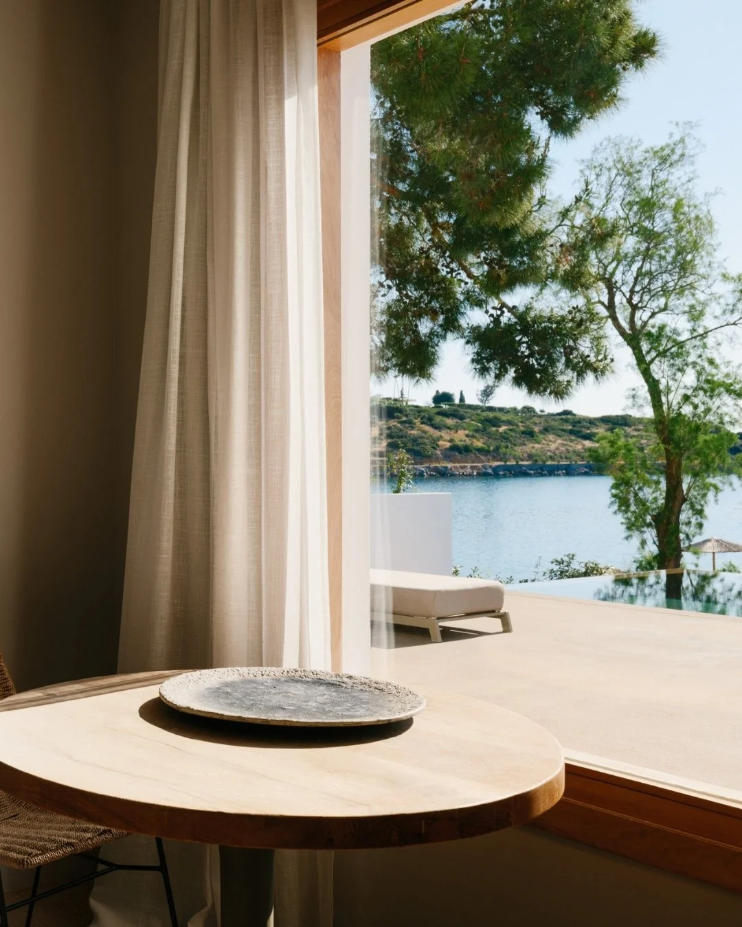Interior view of a room with a round wooden table and a decorative plate, overlooking an outdoor pool and a scenic landscape with water, trees, and hills.