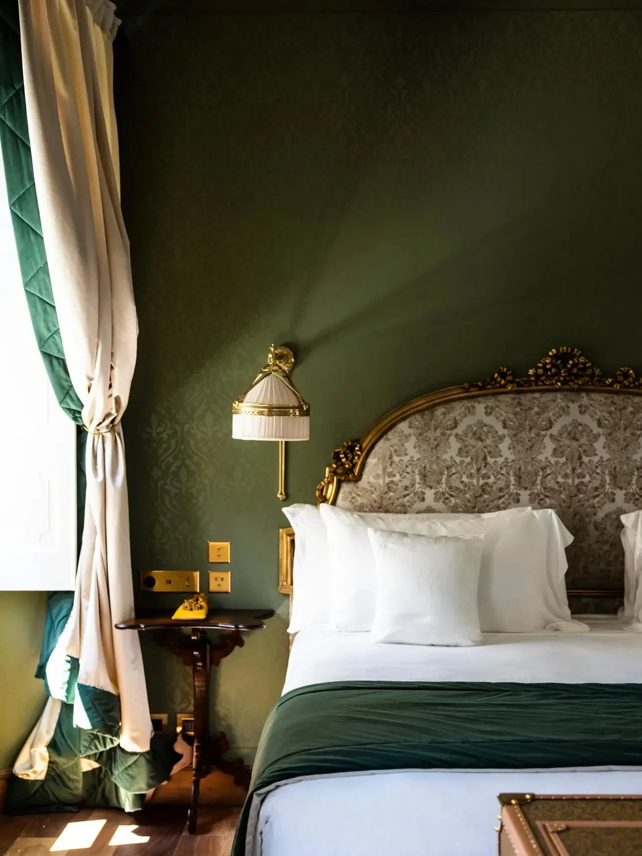 A bedroom with a bed, white pillows, and a patterned headboard. There is a green bed cover at the foot of the bed, a nightstand with a yellow telephone, and a wall-mounted lamp with a pleated shade. Decorative curtains with green and beige colors are gathered on the window, and the wall behind the bed is dark green with a subtle damask pattern.