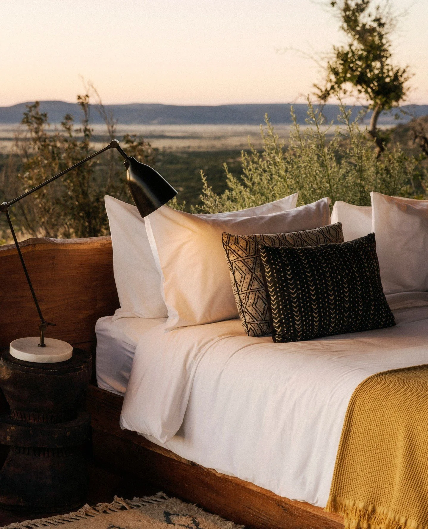 A cozy bedroom with a mountain view in the background, featuring white bedding, patterned pillows, a wooden headboard, a bedside table with a lamp, and a yellow throw blanket.