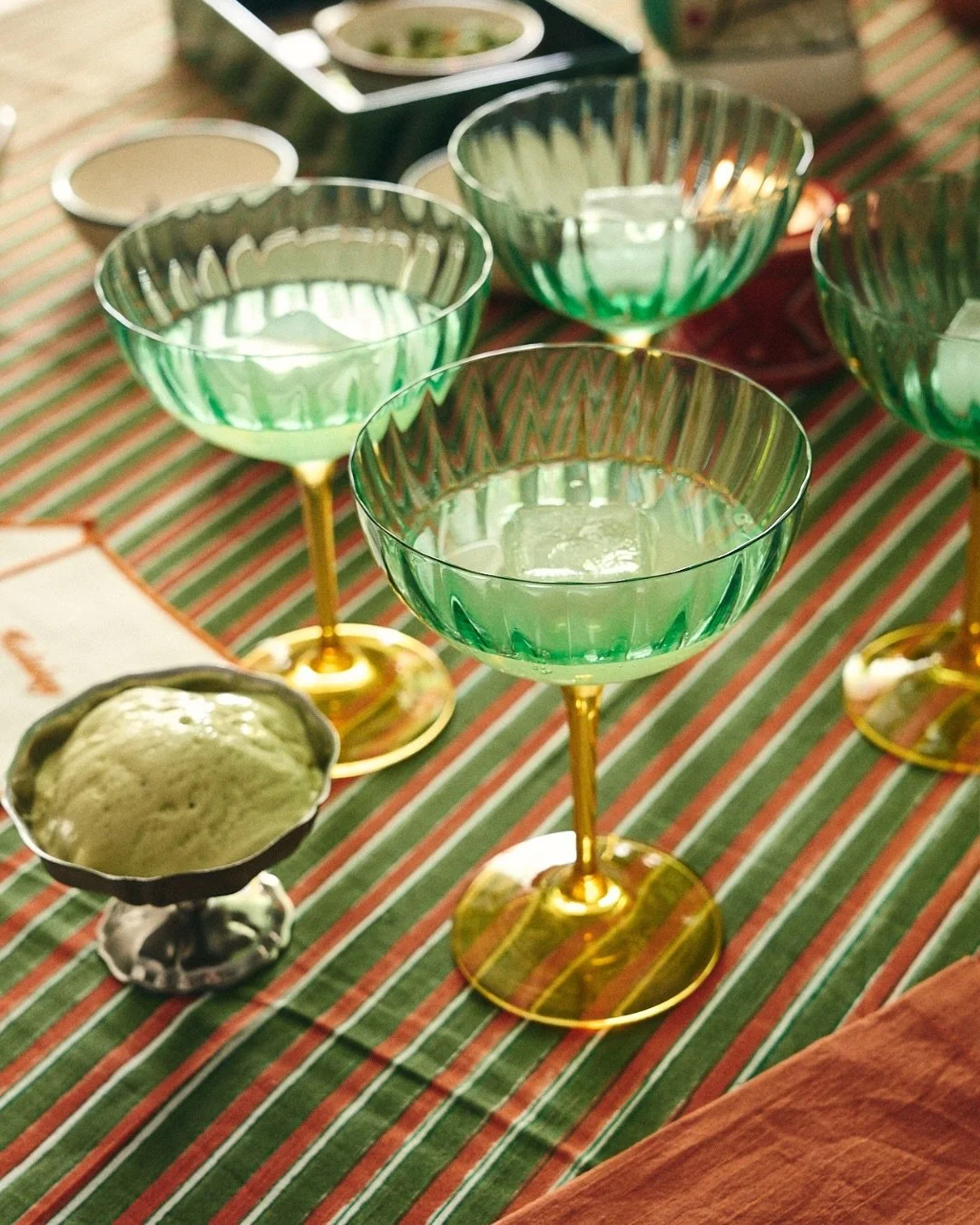 Green cocktail glasses with gold stems on a striped table, with a bowl of green guacamole in the foreground.