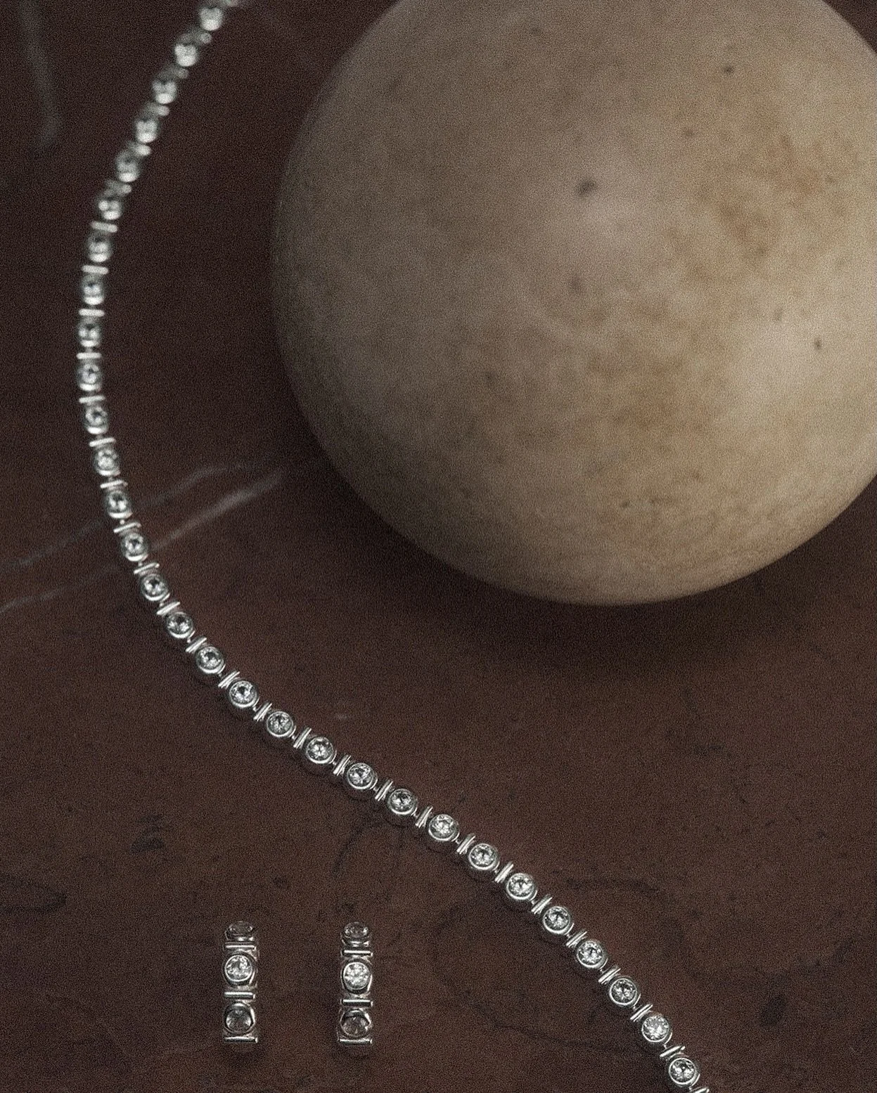 A pearl necklace, a bracelet, and a pair of earrings with diamond-like stones on a dark wooden surface, with an eggshell-colored sphere nearby.