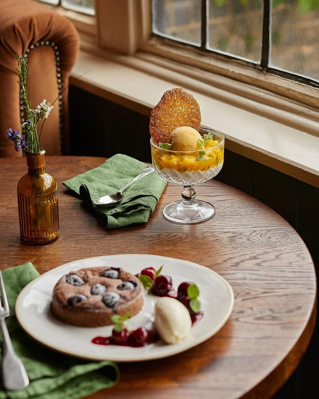 A dessert served in a glass bowl with a scoop of ice cream, a cookie, and garnish, on a wooden table near a window; in front, a plate with a berry tart, cherries, and a scoop of ice cream, and green napkins with a spoon and fork.