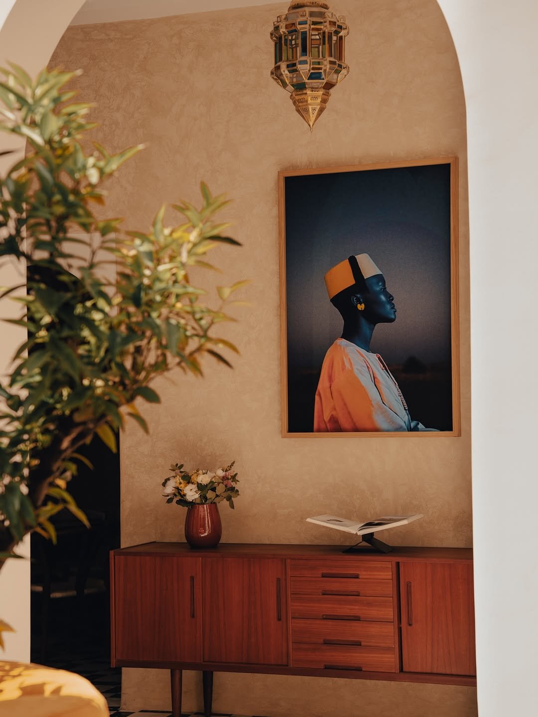 A living room corner with a wooden sideboard, a vase with flowers, an open book on a stand, a framed portrait of a woman wearing a hat, and a Moroccan-style hanging lantern.