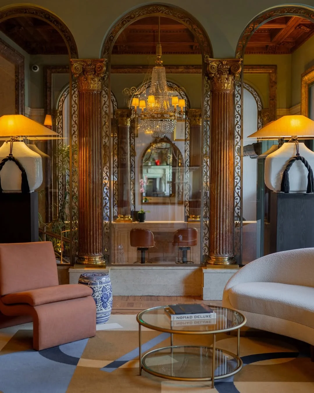 Luxurious living room with ornate columns, a crystal chandelier, and elegant furniture including a pink chair, white sofa, and glass coffee table with books.
