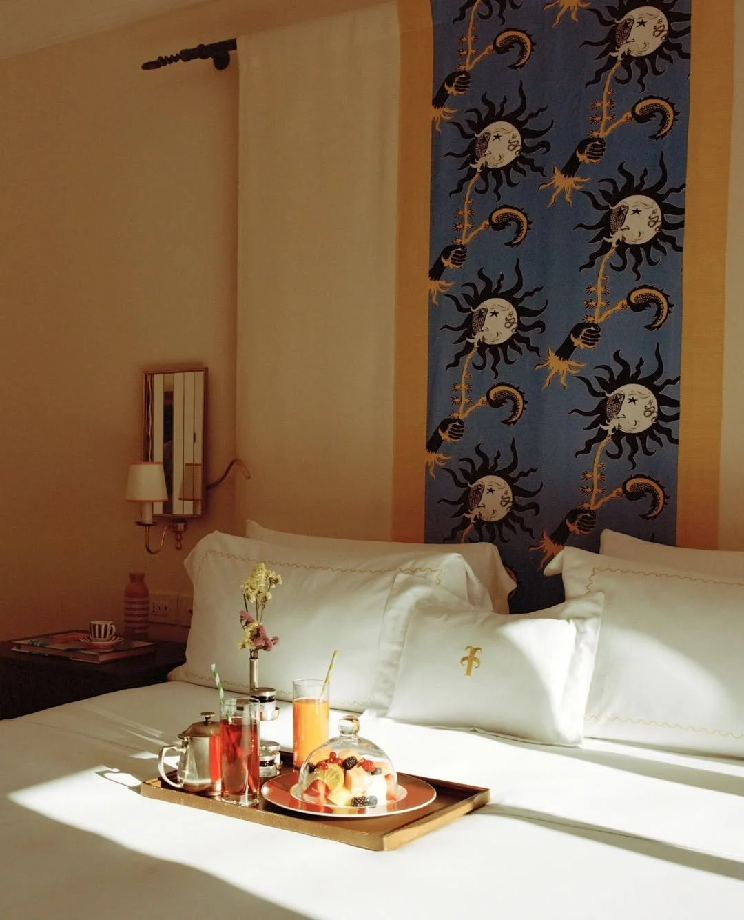 A neatly made bed with white linens and pillows, a tray with drinks and a plate of fruit, and a decorative tapestry featuring sun and moon motifs on the wall behind the bed.
