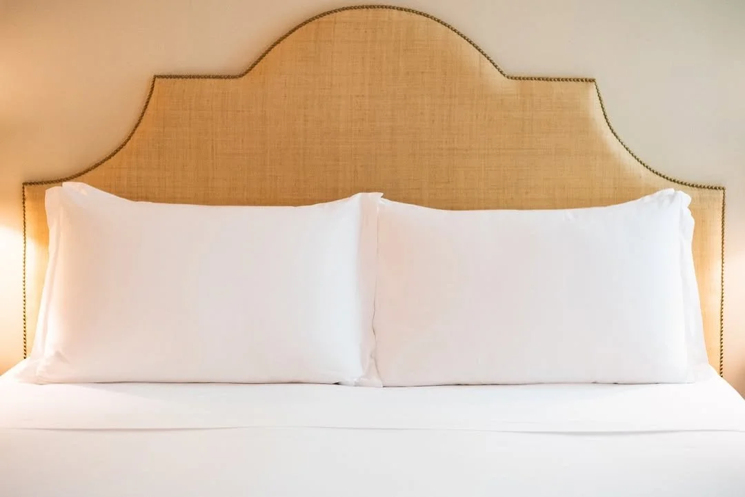 A bed with white pillows and a beige upholstered headboard in a warmly lit room.