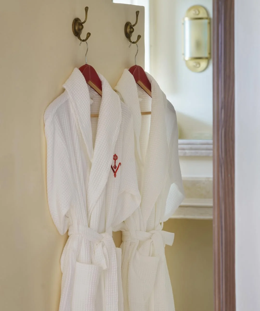 Two white bathrobes hanging on hooks in a bathroom, with a mirror reflecting part of the room in the background.