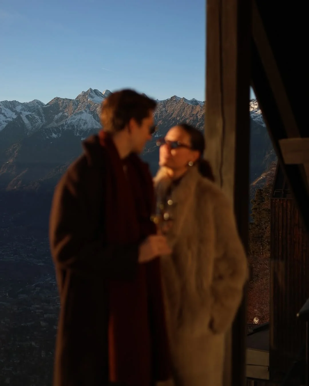 A blurry photo of a man and woman talking, with snow-capped mountains in the background, possibly on a balcony or terrace.