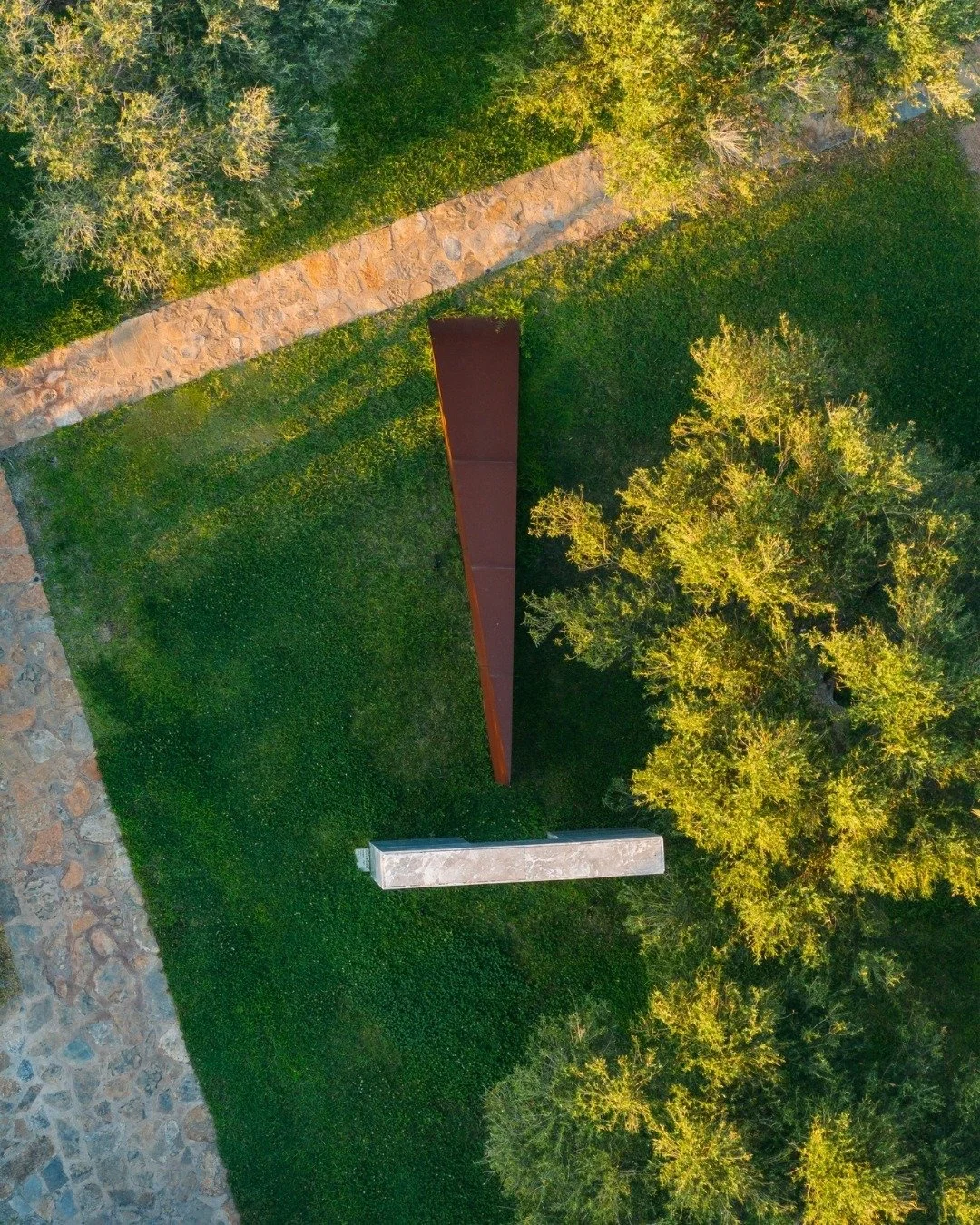 Aerial view of a park with a winding stone path, a large metal bench, a rectangular sculpture, green grassy areas, and trees with yellow-green leaves.