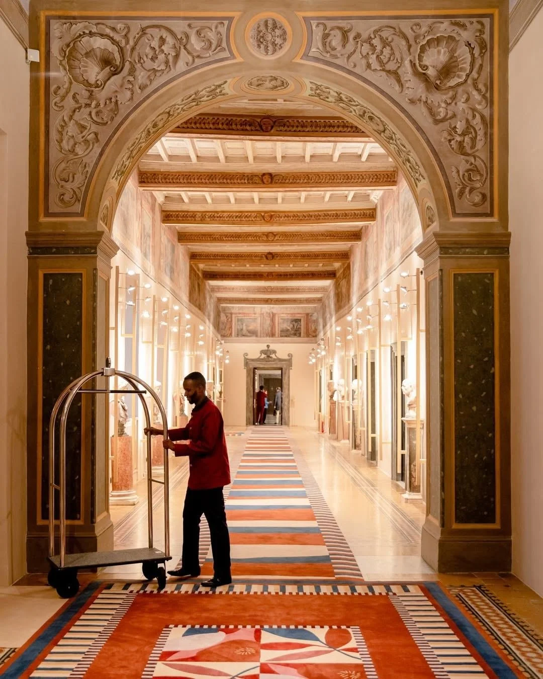 A hotel corridor with a decorative arched ceiling, ornate wall moldings, and a patterned carpet. A bellhop is standing with a luggage cart, and there are people in the background near a doorway.