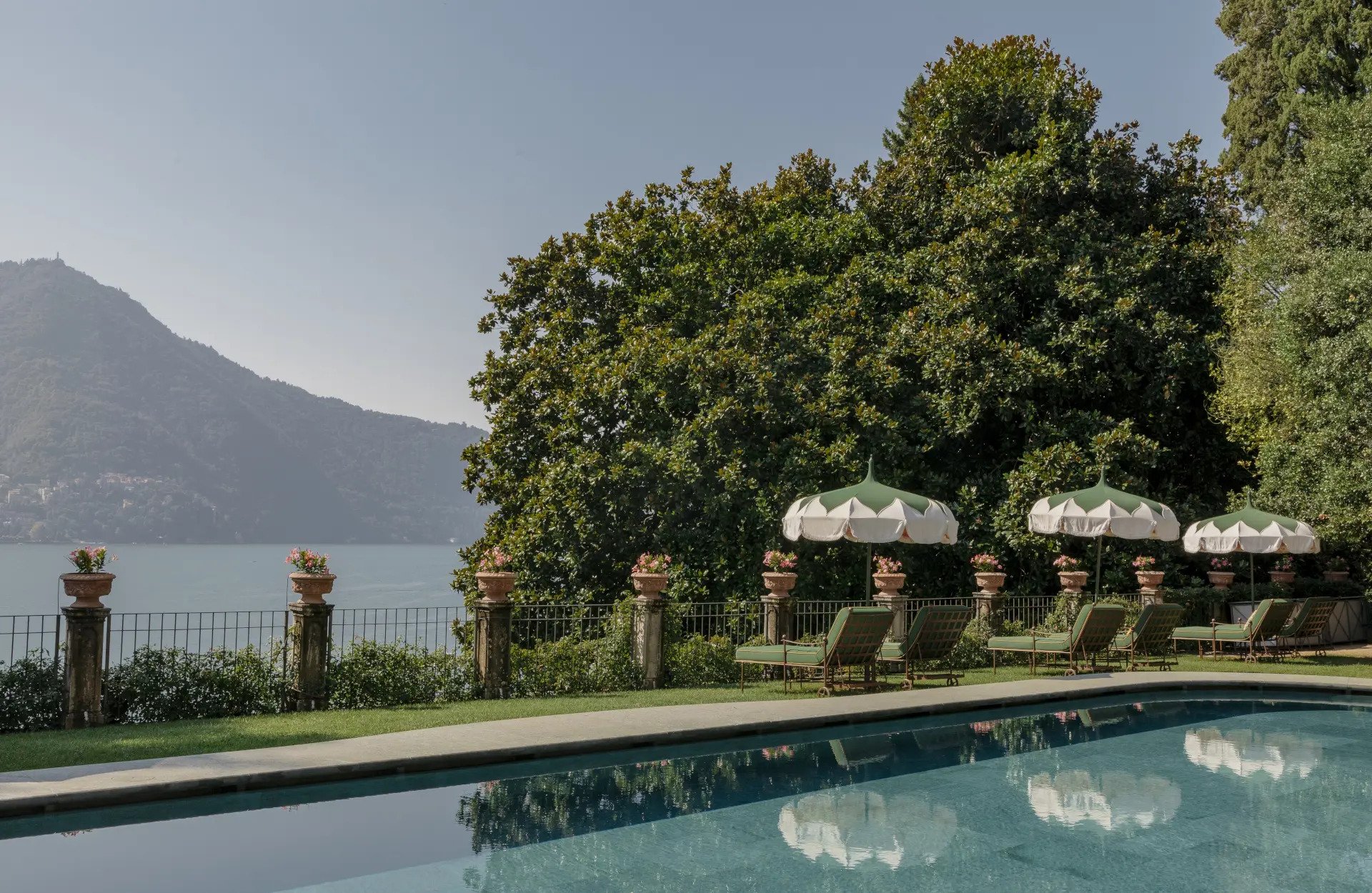 View of a swimming pool with lounge chairs and umbrellas, a stone balustrade with potted flowers, green trees, a lake, and a mountain in the background under a clear sky.