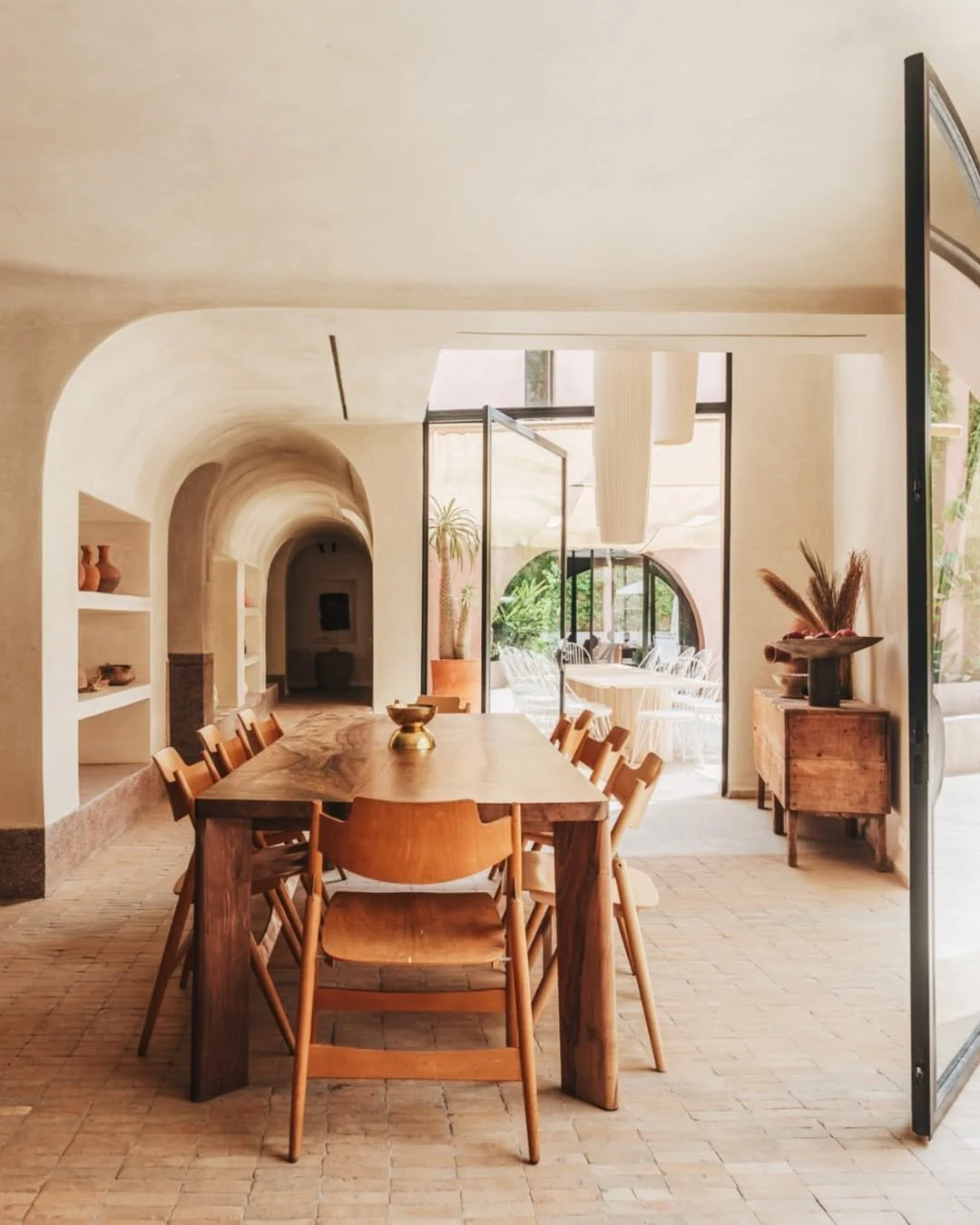 A modern dining room with a wooden table and matching chairs, arched walls, and open glass doors leading to an outdoor patio with white chairs and lush greenery.