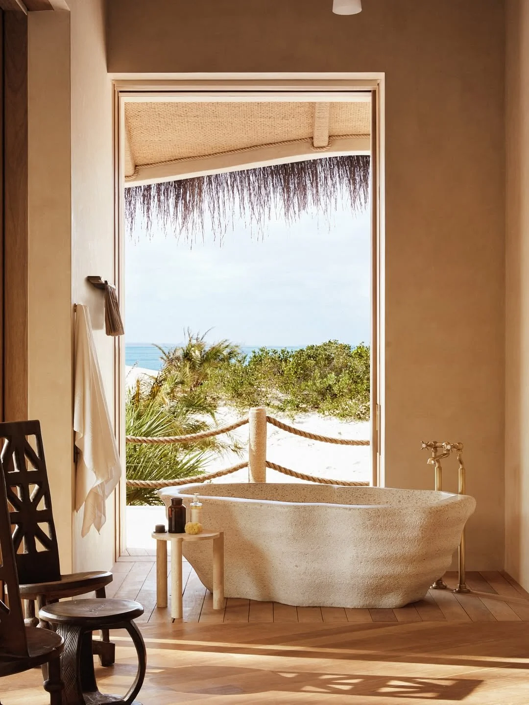 Beach view through open doorway with stone bathtub, small side table with bottles, wooden chairs, and a towel hanging on the wall.