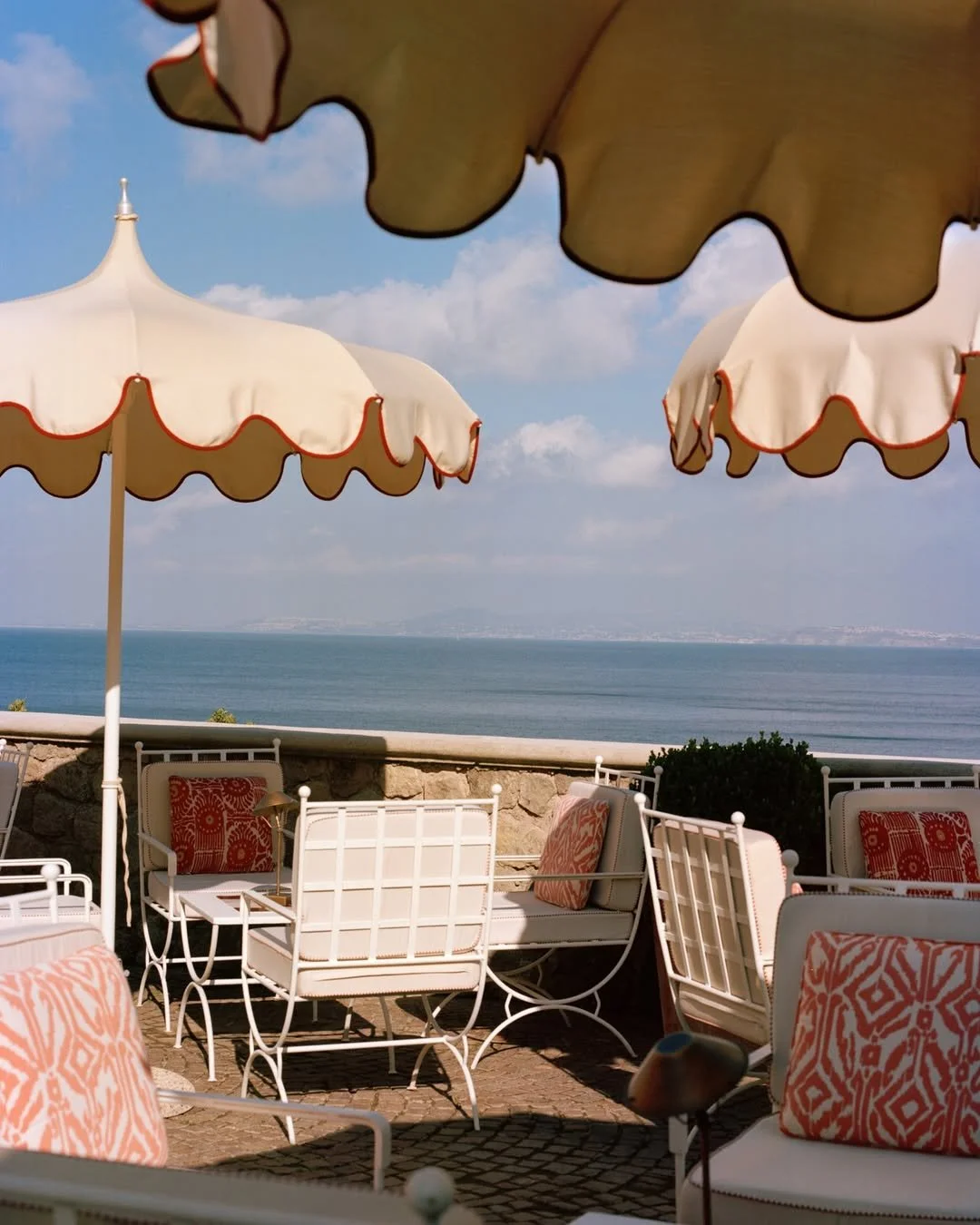 Outdoor patio with white tables and chairs, some with red and white patterned cushions, under large white umbrellas with red trim, overlooking a body of water and a partly cloudy sky.