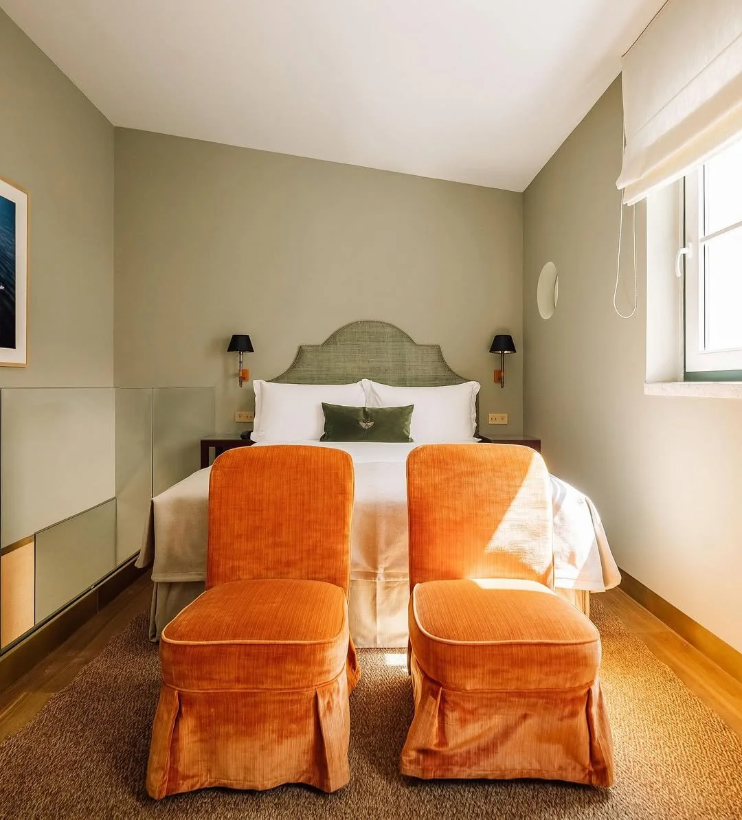 A bedroom with a green headboard, two black wall lamps, an orange velvet chair at the foot of the bed, and a window with a white blind.
