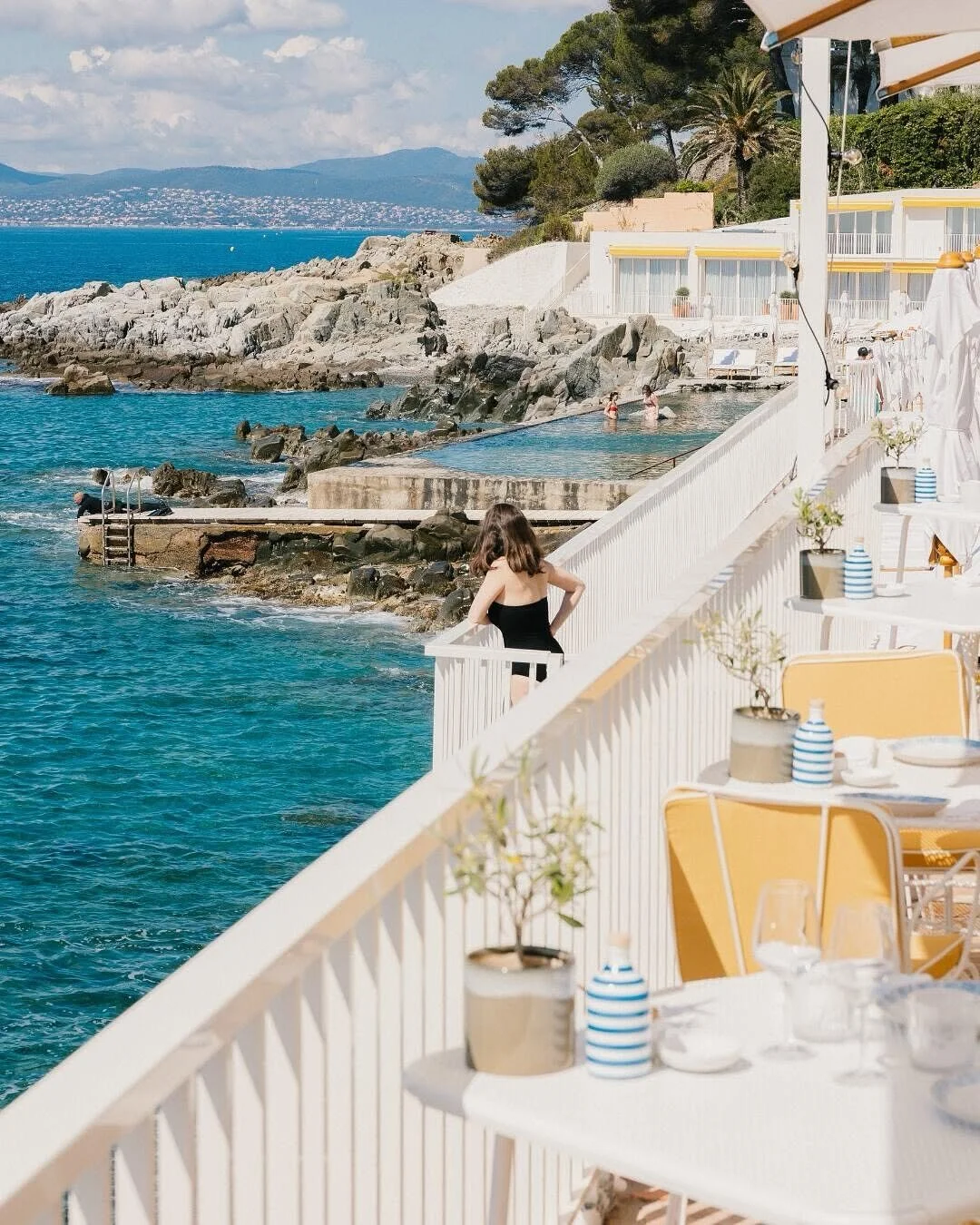 A woman in a black swimsuit stands on a white balcony overlooking the ocean, with rocks and other people swimming nearby, and a beachside restaurant with yellow chairs and striped vases in the foreground.