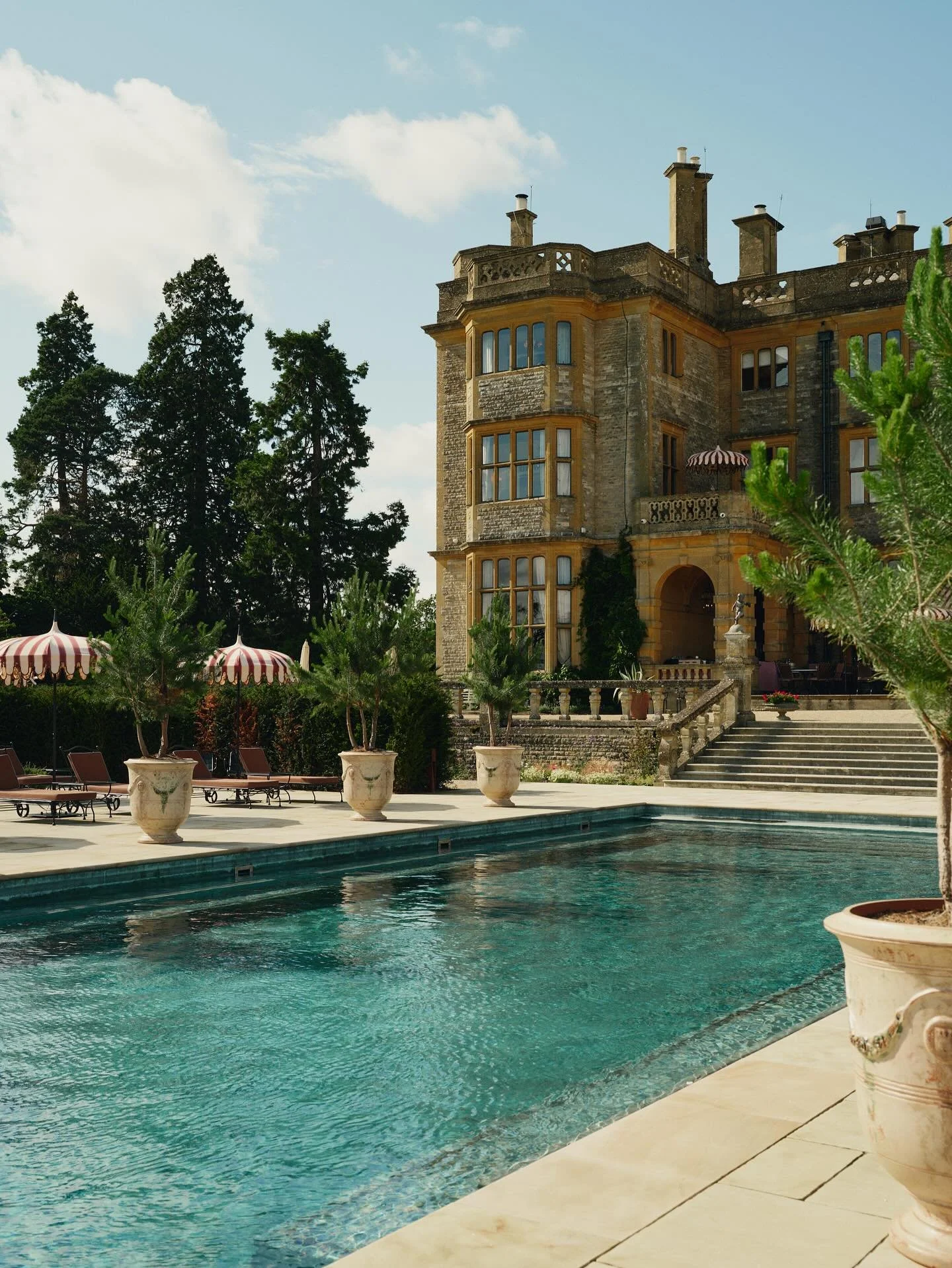 Luxury mansion with a large swimming pool in the foreground, surrounded by potted plants and lounge chairs with striped umbrellas, and a historic stone building with multiple chimneys and large windows in the background, under a partly cloudy sky.