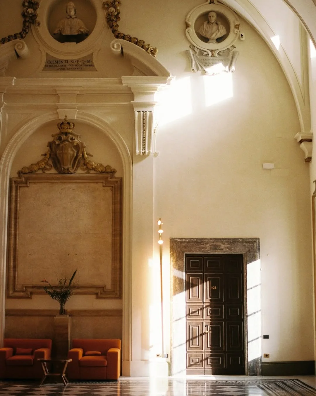Ornate hotel lobby with high ceiling, decorative moldings, portraits, and a large wooden door with the number 108.