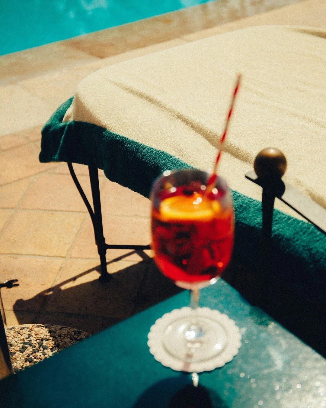 A glass of red cocktail with a lemon slice and a striped straw on a white doily, placed on a teal ledge outdoors near a pool.