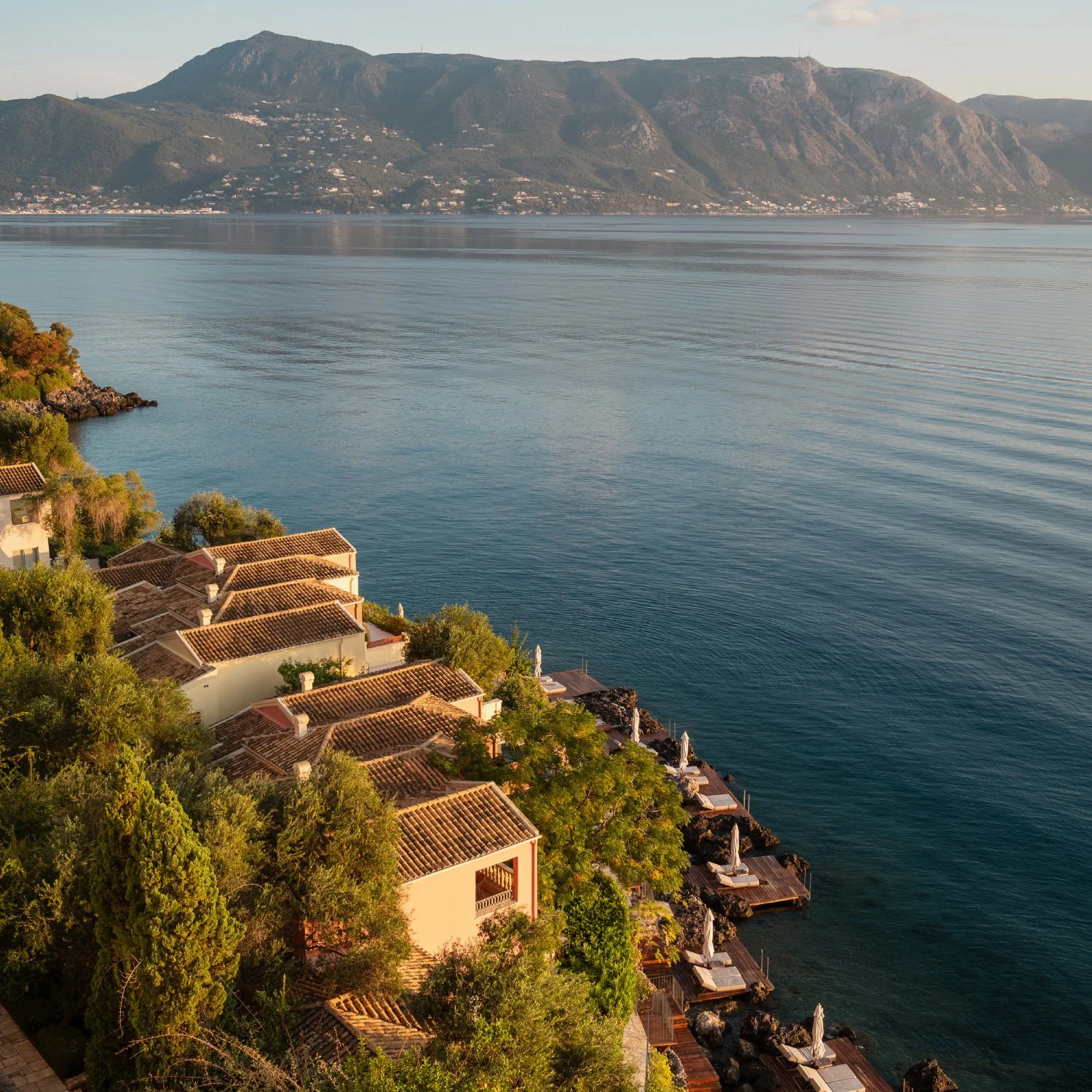 A scenic view of a lakeside village with terracotta-roofed houses surrounded by lush green trees. The calm water extends to distant mountains under a clear sky.