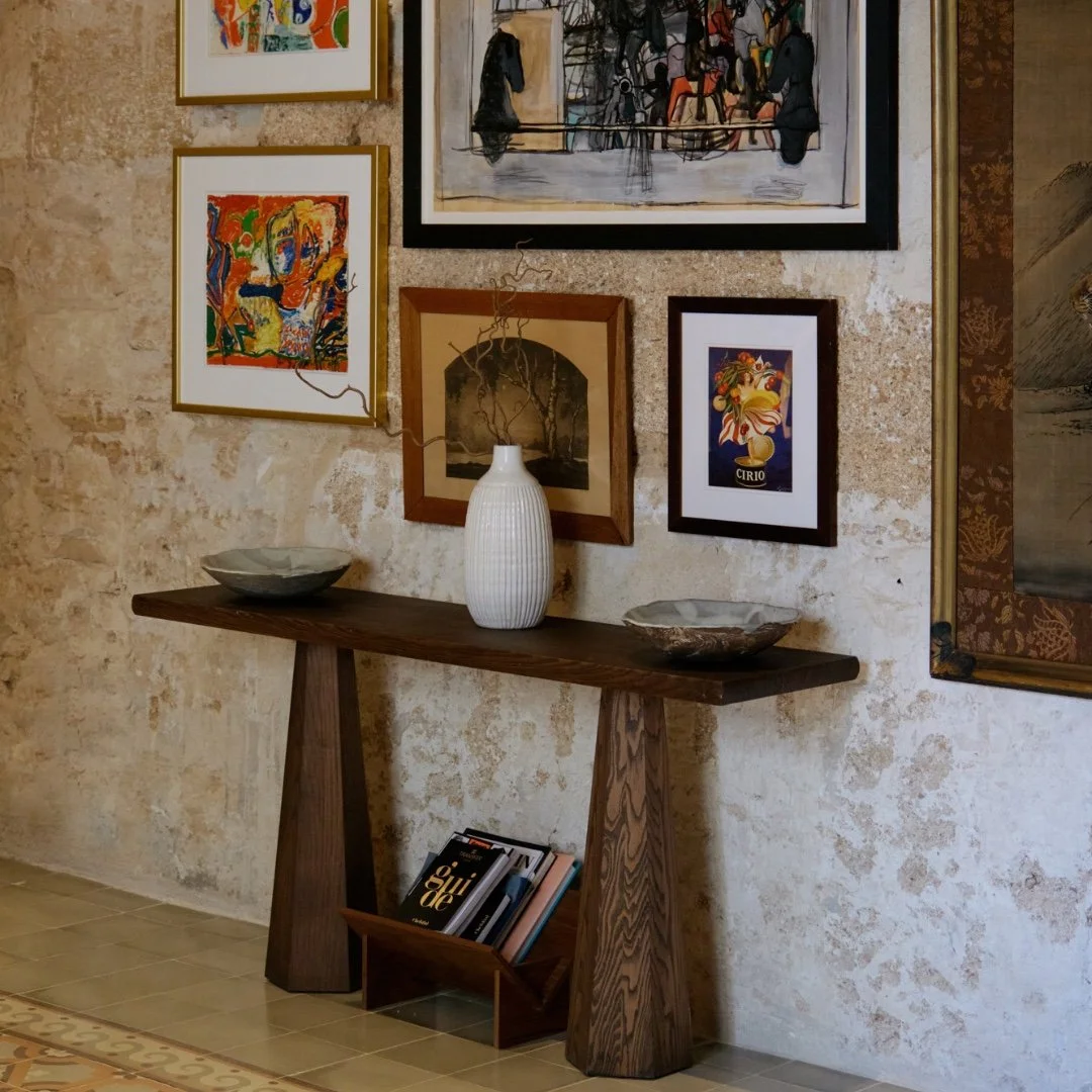 A wall in an art gallery with various framed artworks, a rustic wooden console table with a white ceramic vase and two decorative bowls, and a magazine holder filled with books on a tiled floor.