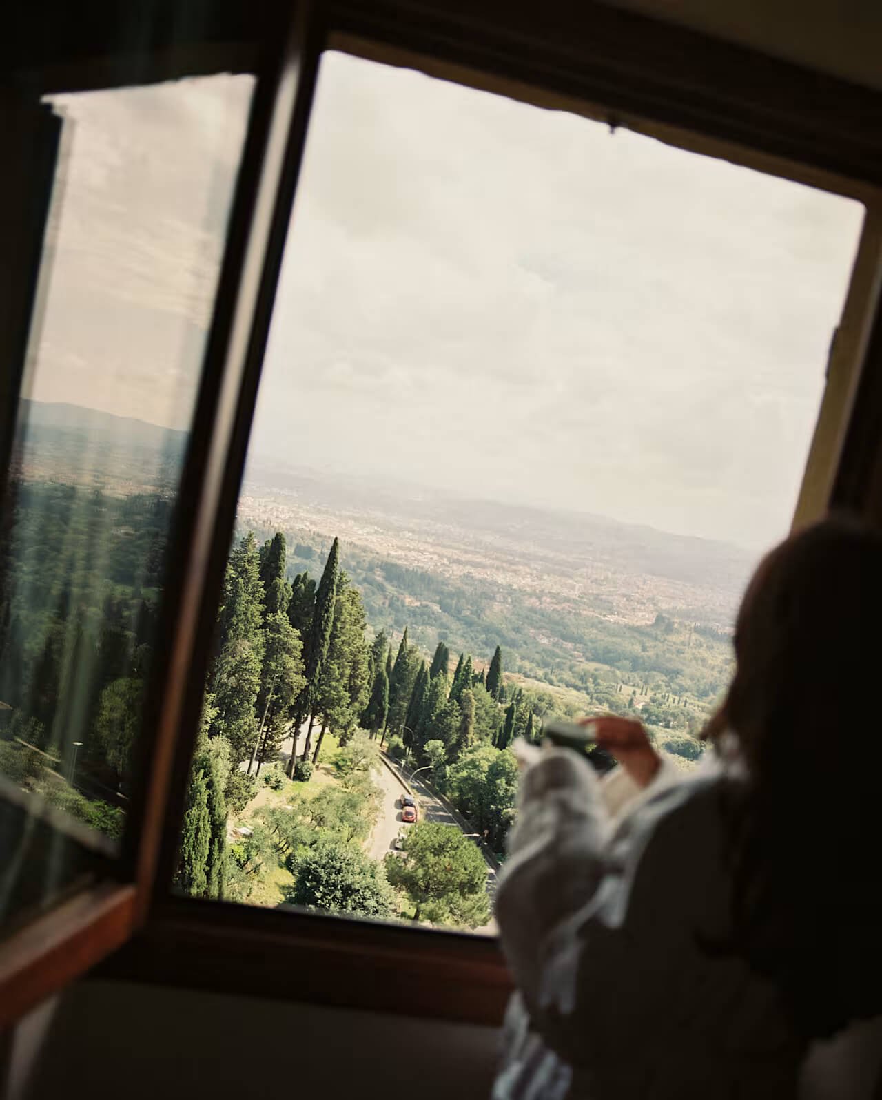 A person looking out a window at a scenic view of green trees, a winding road, and a city landscape in the distance with cloudy sky.