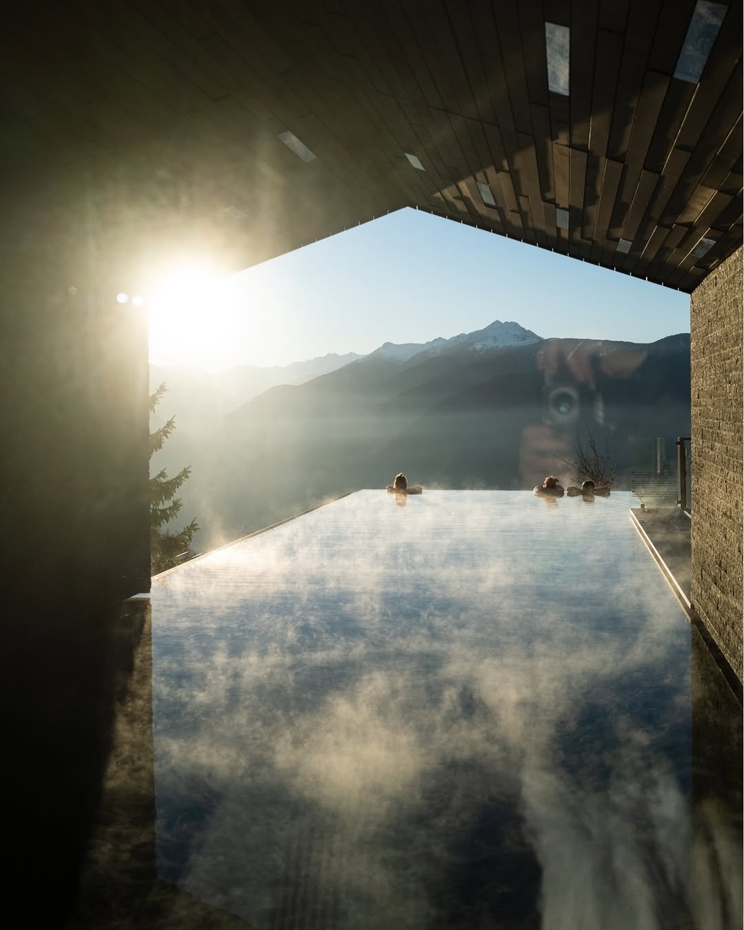 People relaxing in hot spring pool with mountain view and snow-capped peaks, sunny weather, wood ceiling, and stone walls.